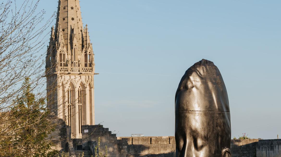 La statue en bronze de "Lou" est posée sur l'herbe. Il s'agit d'un visage de jeune fille. Dans un grand ciel bleu se découpe l'église Saint-Pierre en arrière-plan.