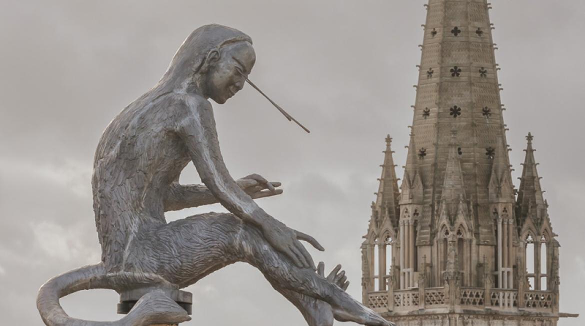Zoom sur le singe à tête d'homme, juché sur son mât en bois, devant la cathédrale Saint Pierre. Le temps est nuageux.