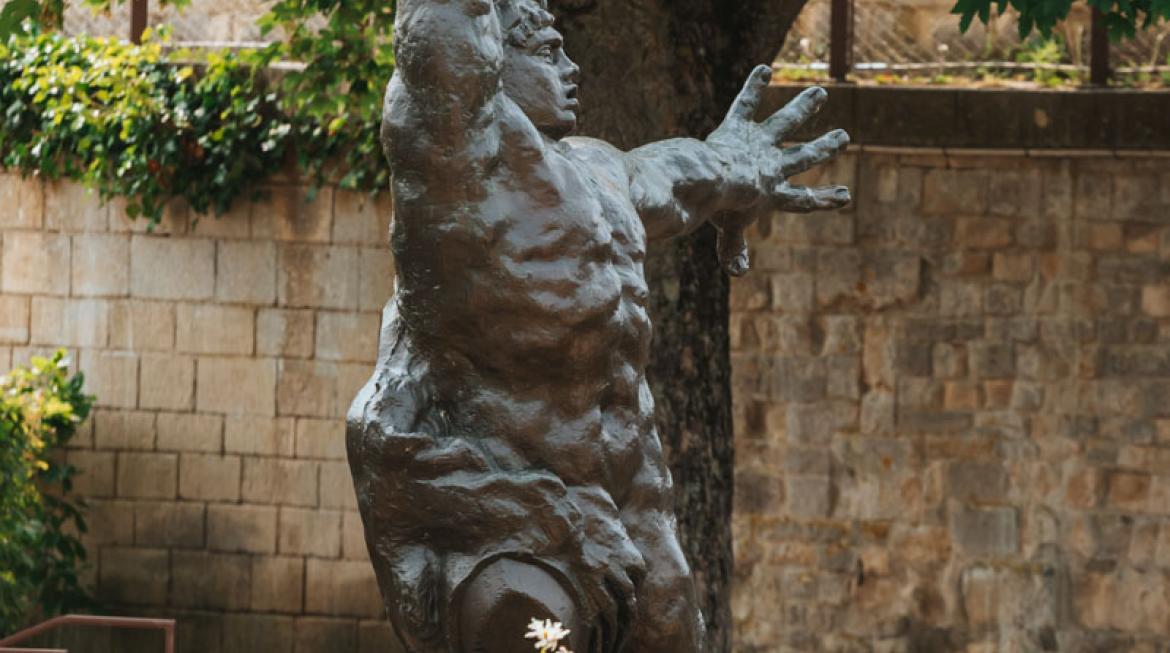La statue du Grand Guerrier de Bourdelle trône au centre de l'image. Elle est sur un socle, au-dessus de marguerites en fleur.