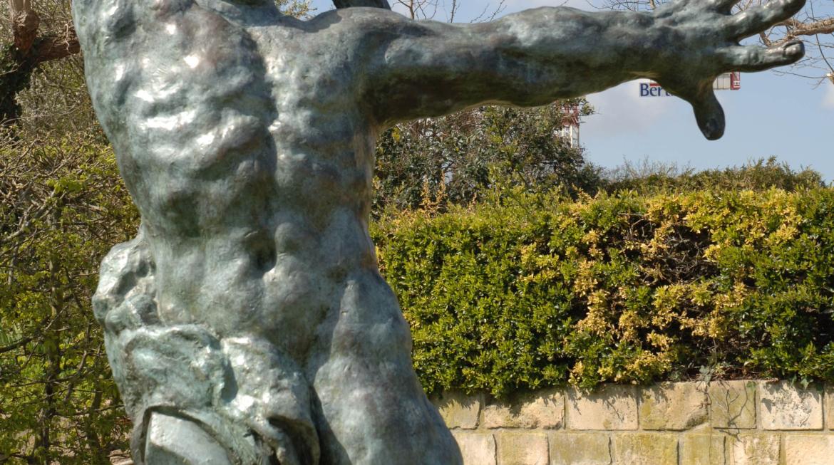 La statue du Grand Guerrier de Bourdelle trône au centre de l'image. Elle est sur un socle, au-dessus d'une étendue de pelouse verte. Il y a un grand ciel bleu.