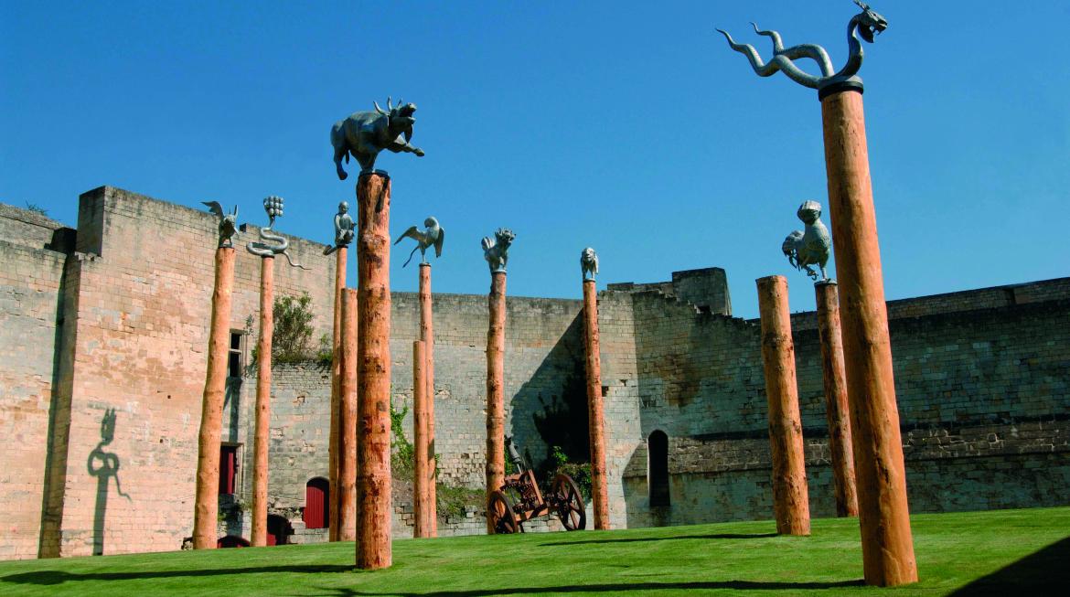 Les sculptures d'animaux juchées sur des mâts se découpent dans un grand ciel bleu. Derrière, la muraille du château se tient fière, et à son pied de la pelouse verte tapisse le sol du parc.